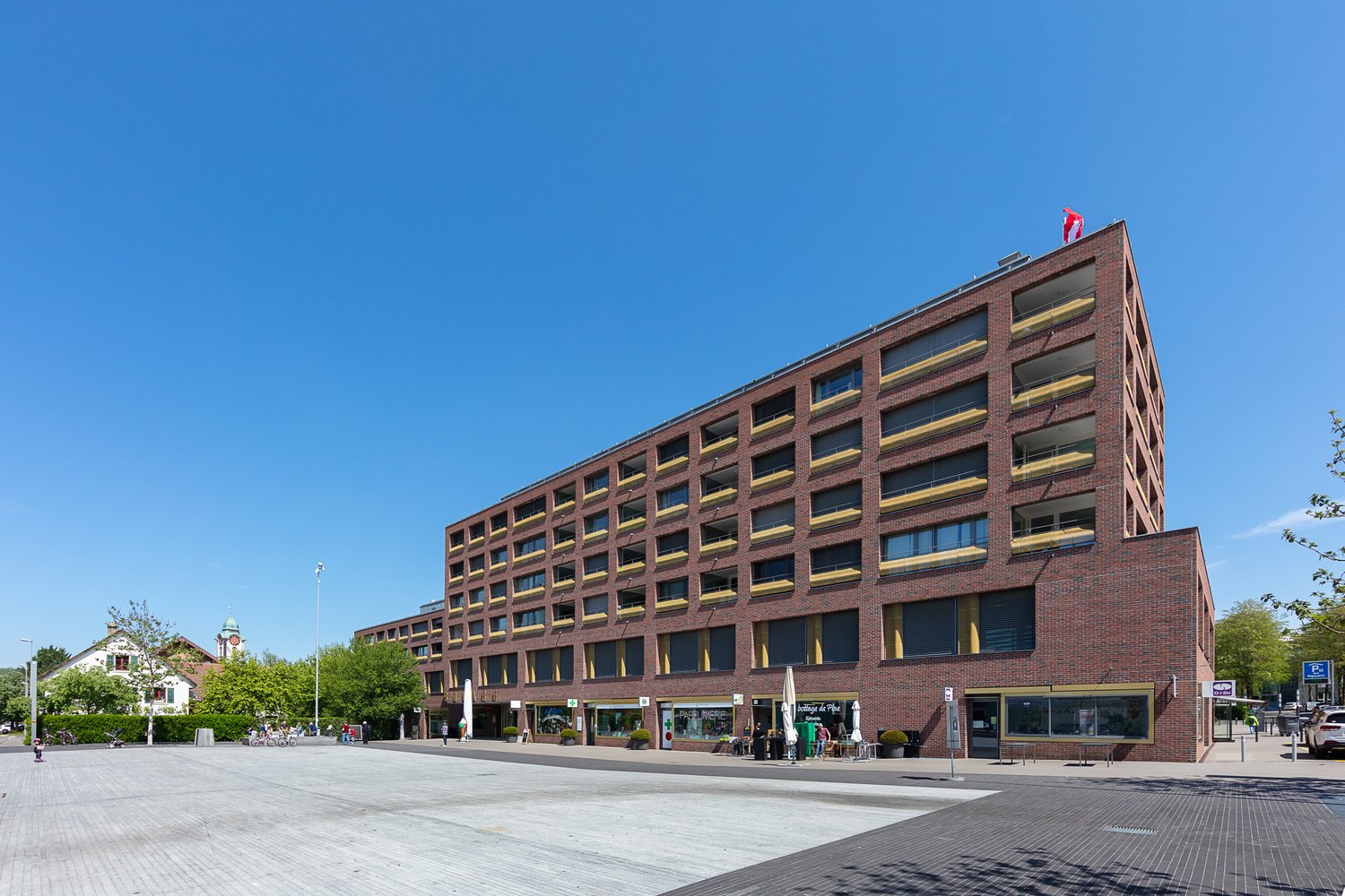 5 story brick building, several balconies, flag on top, storefronts, parking lot in front