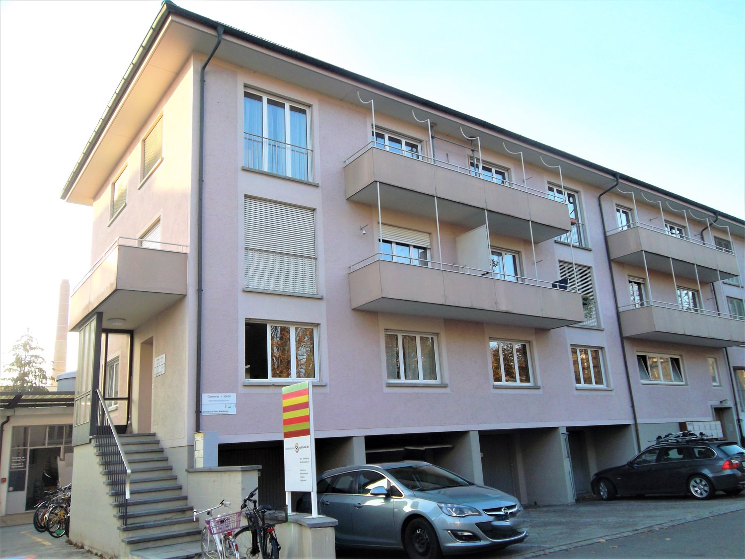 3-story apartment building with balconies, parking spaces in front, and a sign indicating the building's name or address