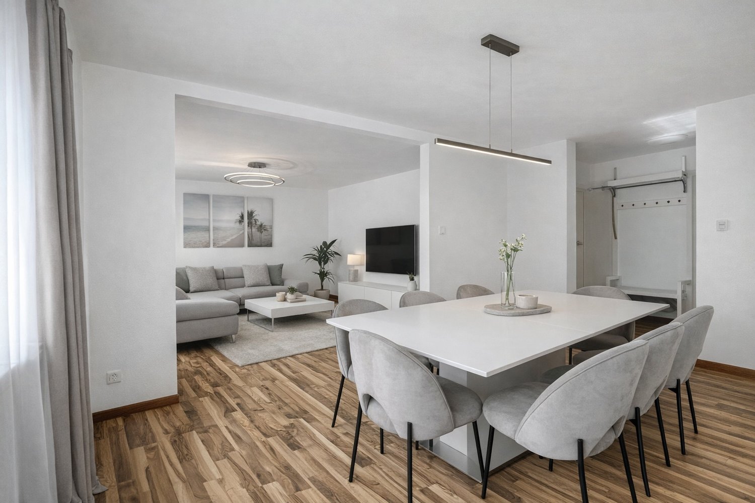 Dining room with a large white table, gray chairs, wooden floor, white walls, a gray couch, a coffee table, a television, and a plant.