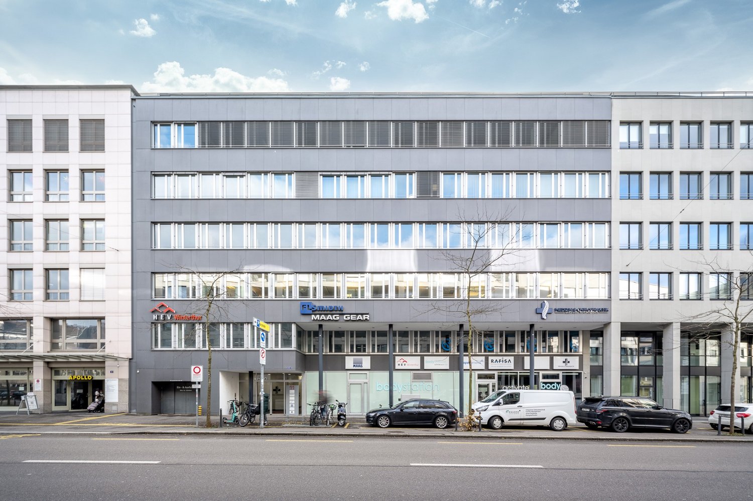 Modern office building with glass windows, multiple floors, entrance with glass doors, bicycles parked outside, cars parked in front