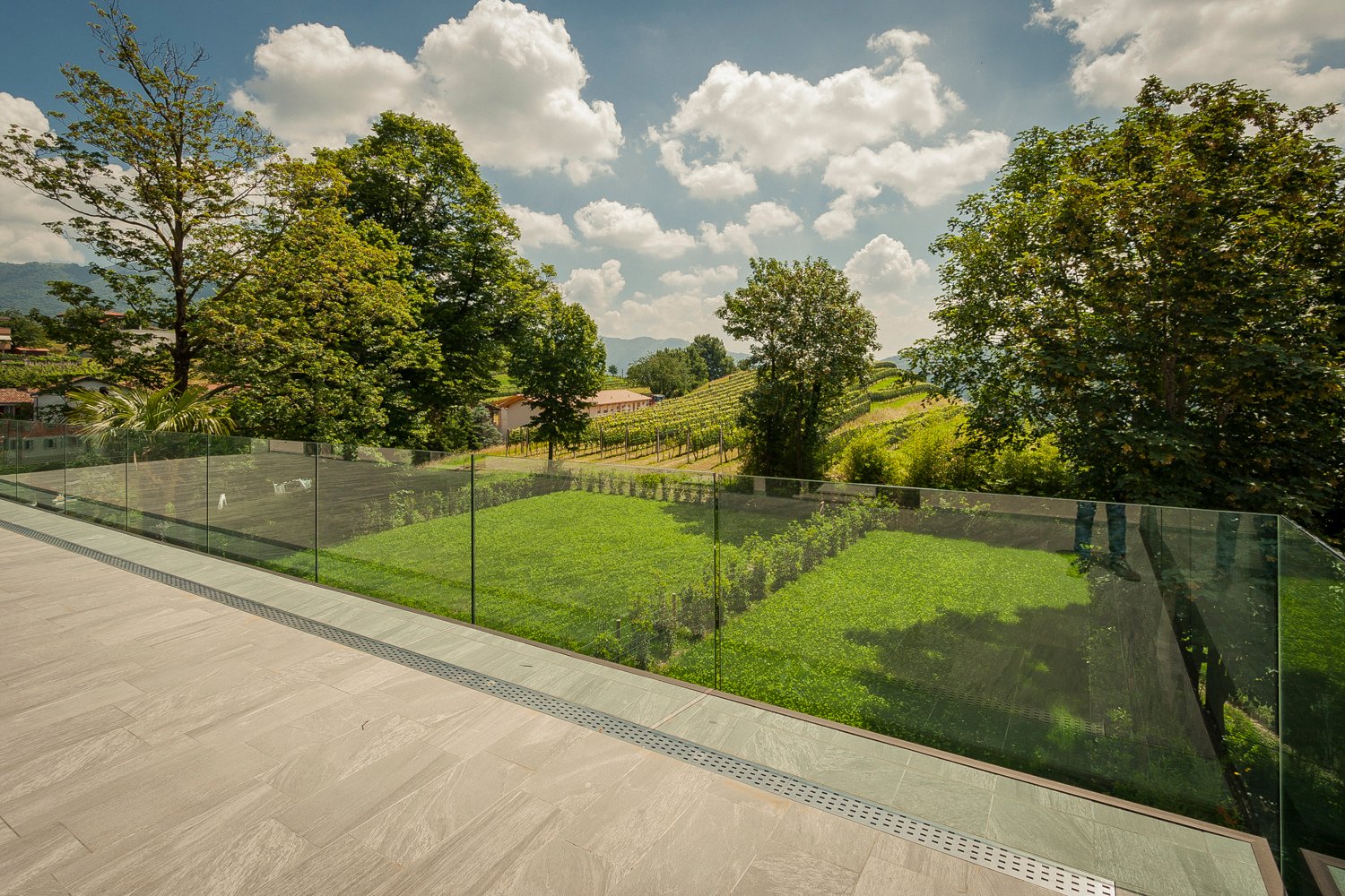 Large terrace with glass railings and green lawn, mountains and buildings in the background.