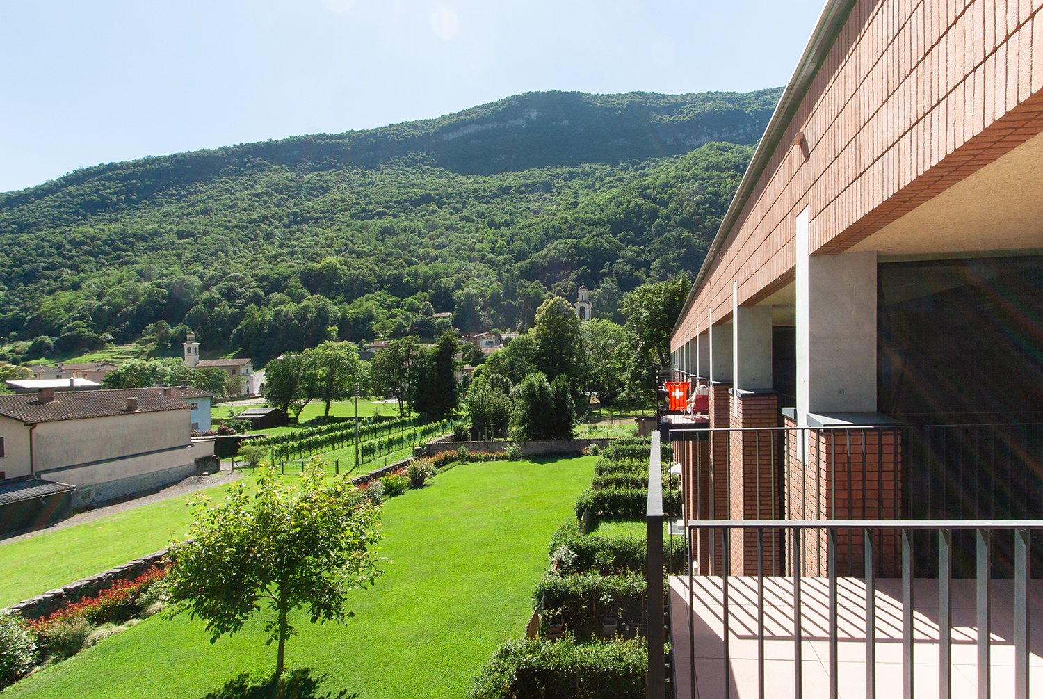 A multi-story building with a balcony overlooking a lush, green landscape. The building is surrounded by a well-manicured garden with trees, shrubs, and a grassy lawn. In the distance, there are hills covered in dense vegetation.