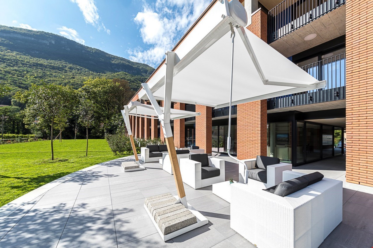 Modern multi-story building with brick facade, large windows, and a covered outdoor seating area with white umbrellas and furniture. The building is surrounded by lush greenery and mountains in the background.