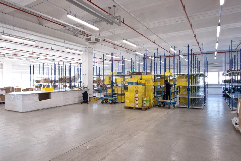 Large industrial warehouse space with shelving units, storage boxes, and a reception desk. The space has high ceilings, concrete floors, and overhead lighting.
