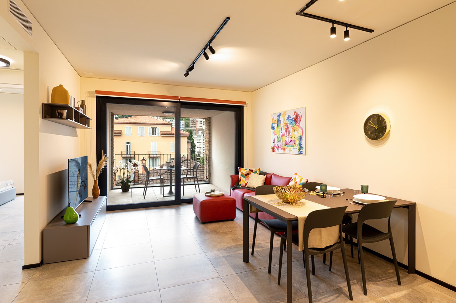 Modern open-plan living area, dining area with glass door leading to balcony, beige flooring, dining table, chairs, sofa, TV, painting, wall clock, ottoman, green vase, decorative items, view from balcony