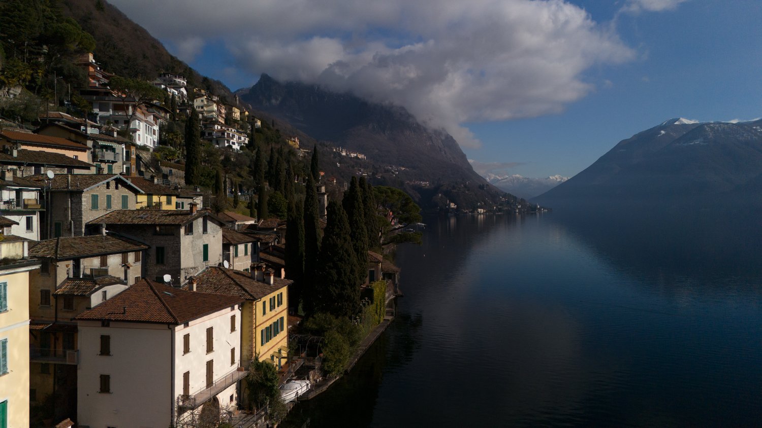 The scene shows a mountain village beside a body of water, surrounded by hills, and features multiple buildings, lush greenery, and a serene atmosphere.