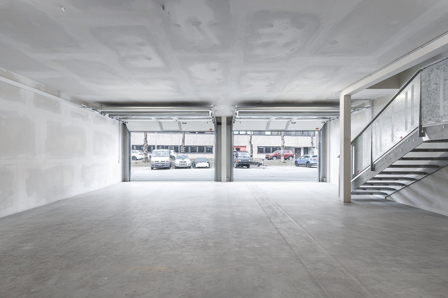 Empty garage with two open doors, concrete floor, white walls, metal staircase
