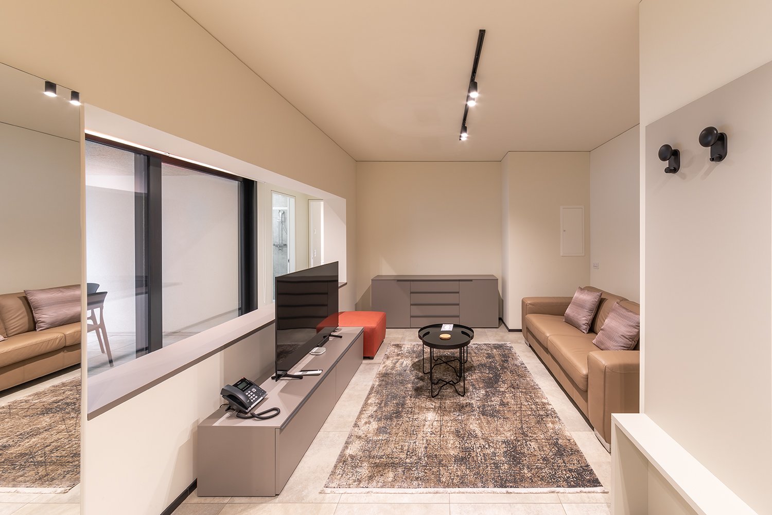 modern living room with beige walls, large windows, beige couch, TV, coffee table, rug, telephone