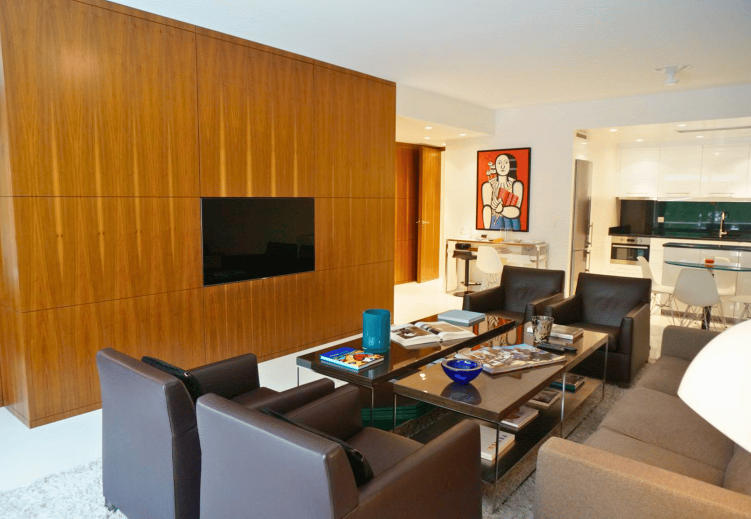 Living room with modern interior, wooden wall, couch, chair, coffee table, tv, artwork, kitchen and dining area visible in background