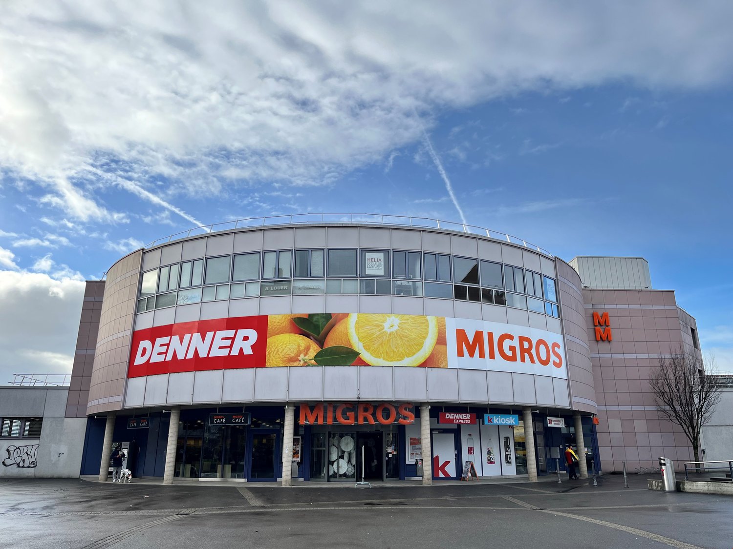 a modern, commercial building with storefronts, glass windows, and a curved facade. There are large advertisement boards with pictures of oranges on it.