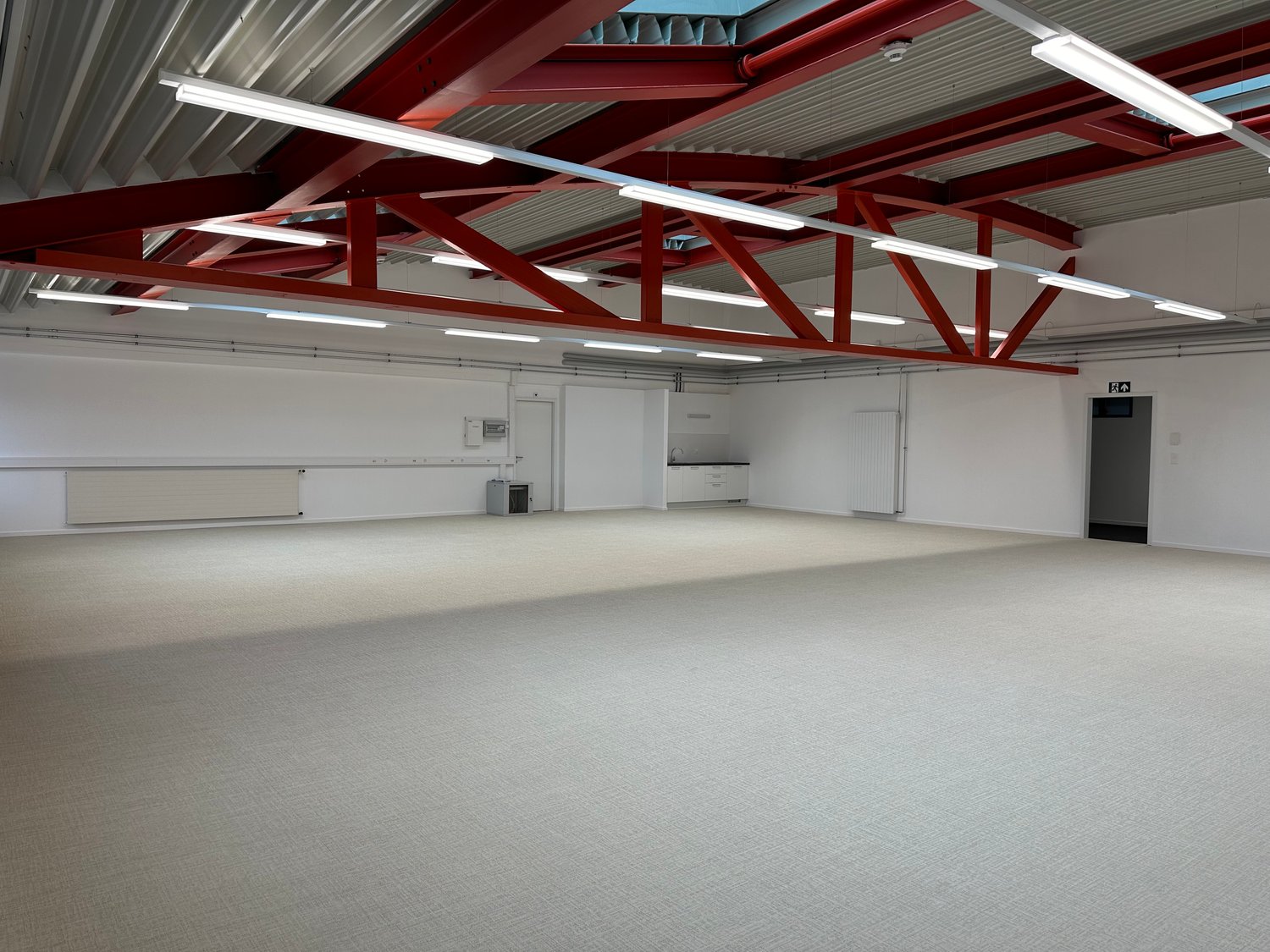 Large open space with high ceilings, red metal beams, and white walls. The room appears to be a warehouse or industrial space with minimal furnishings.