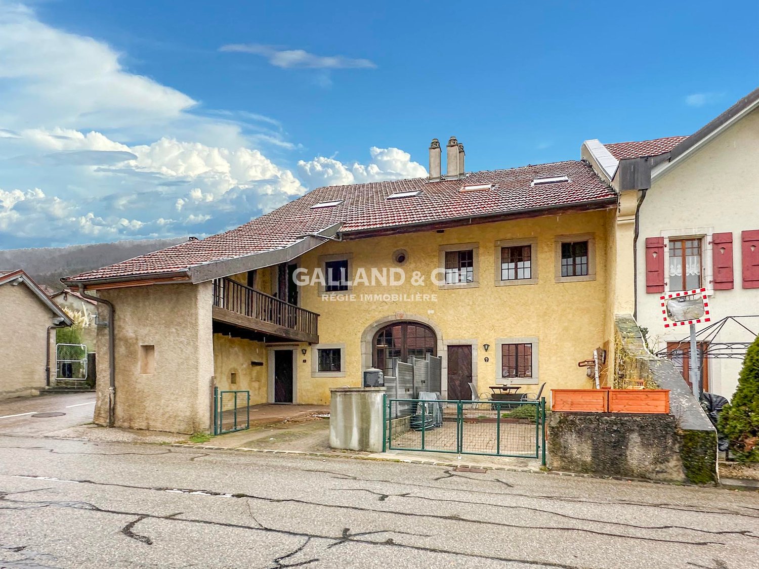 two-story house, yellow facade, tiled roof, wooden balcony, chimneys, fenced garden, driveway, green gates