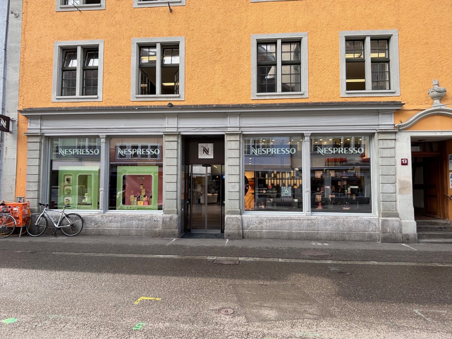 A building with orange walls, four windows, and a glass door with the Nespresso logo. A bicycle is parked in front of the building on the sidewalk.