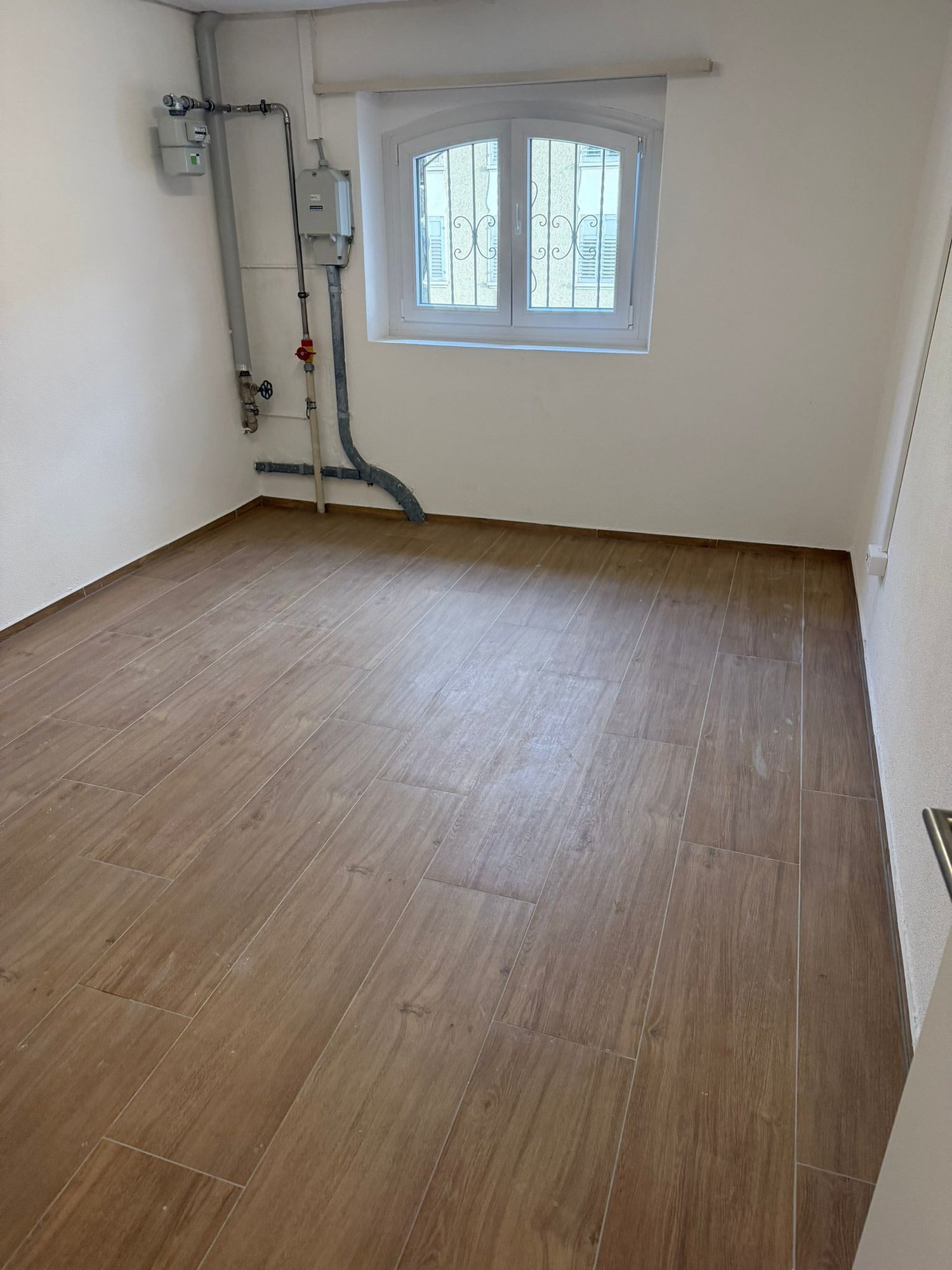 empty room, wooden floor, white walls, window with metal grill, pipes on the wall