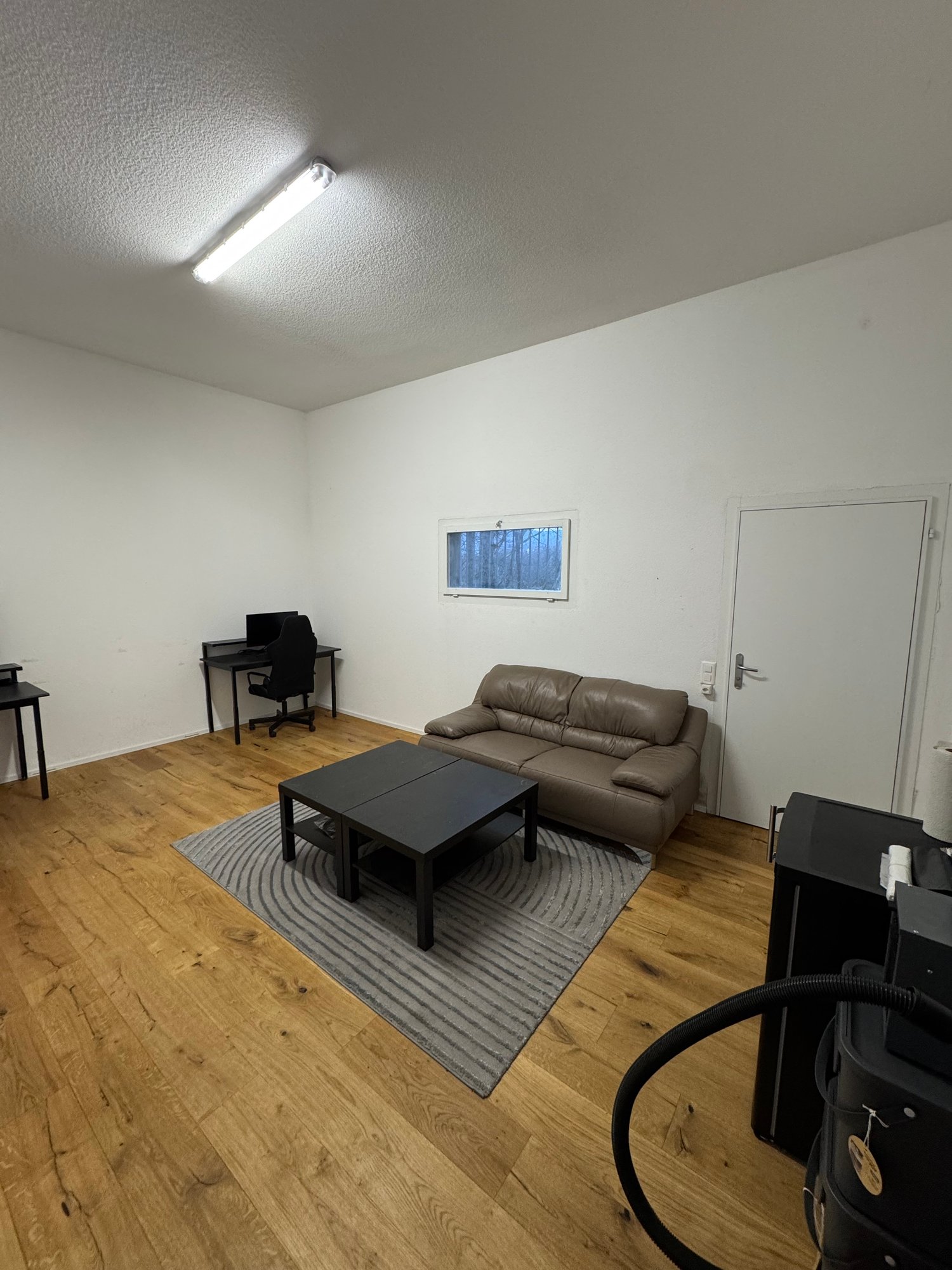 Empty room with wooden floor, white walls, a couch, a desk, a chair, a window, and a rug