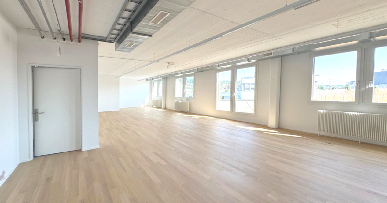large room, hardwood floors, white walls, multiple windows, door at left, radiators