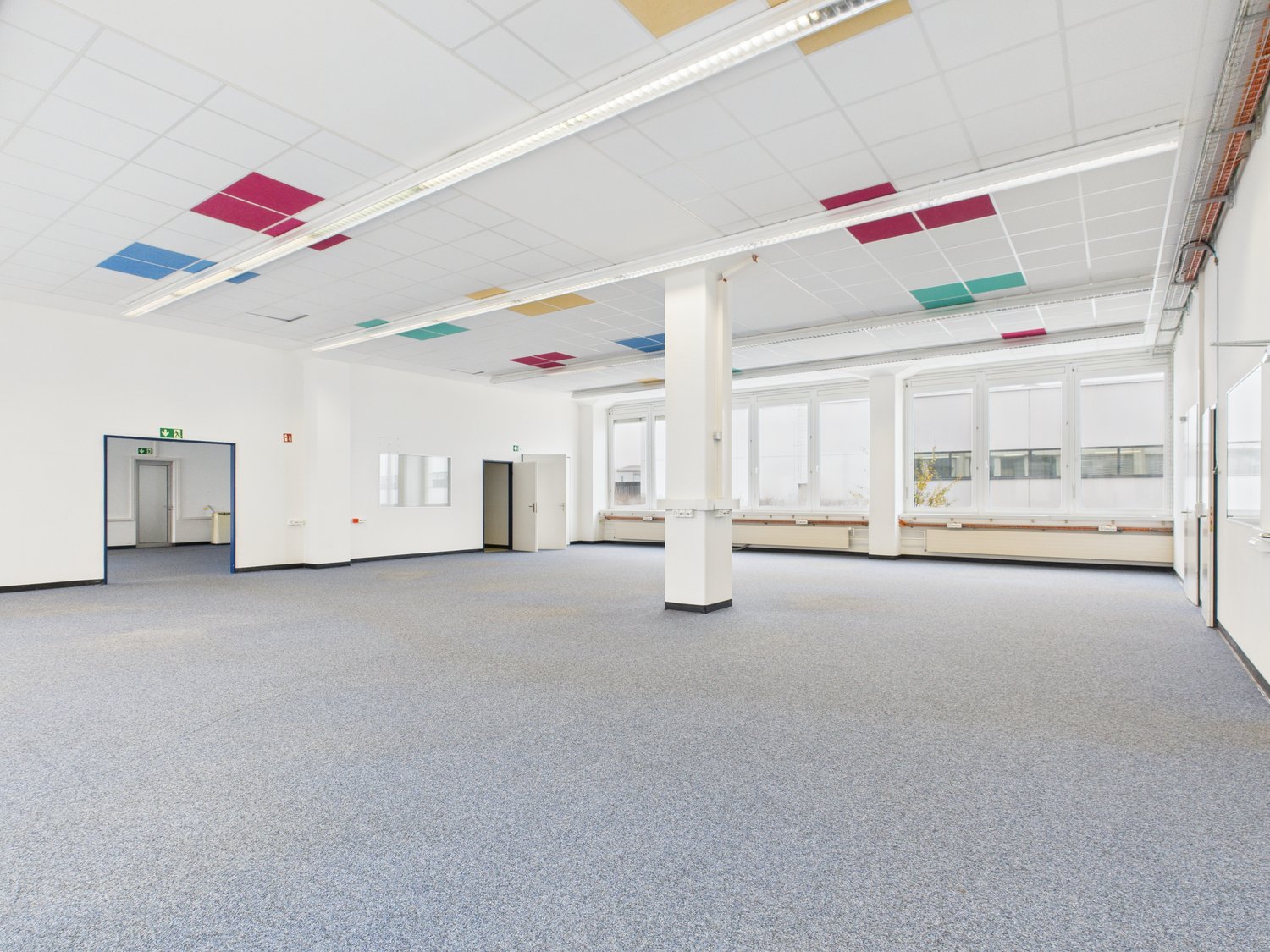 Empty room, white walls, gray carpet, multiple large windows, white column in the center, square ceiling tiles