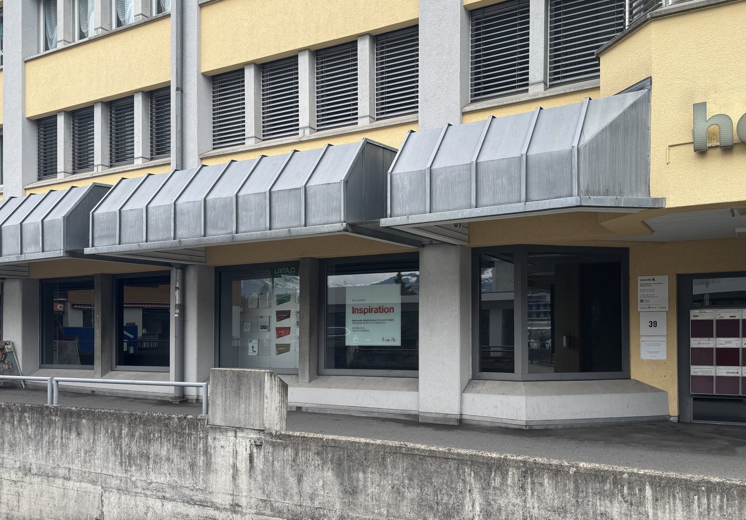 2 story building, yellow wall, grey roof, entrance with metal railings, glass windows, large awnings