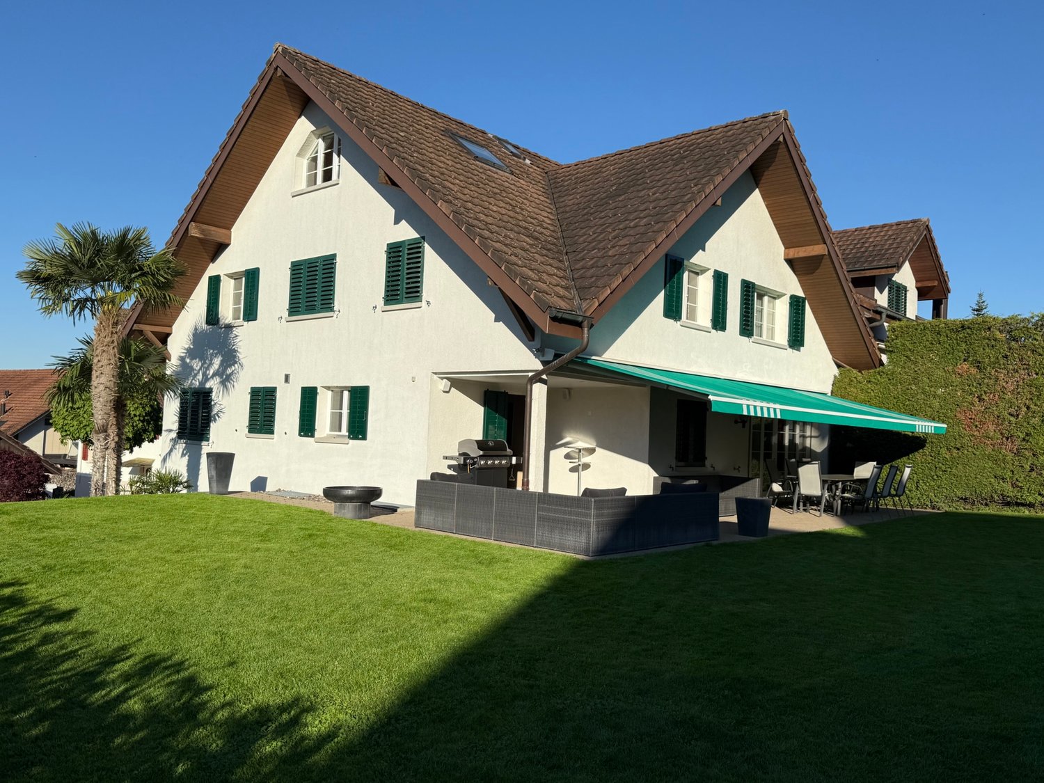 2-story house with a brown tiled roof, white exterior walls, and green shutters. The house has a covered patio or balcony area with a retractable awning. There is a garage and additional parking space on the property. The house is surrounded by a well-man