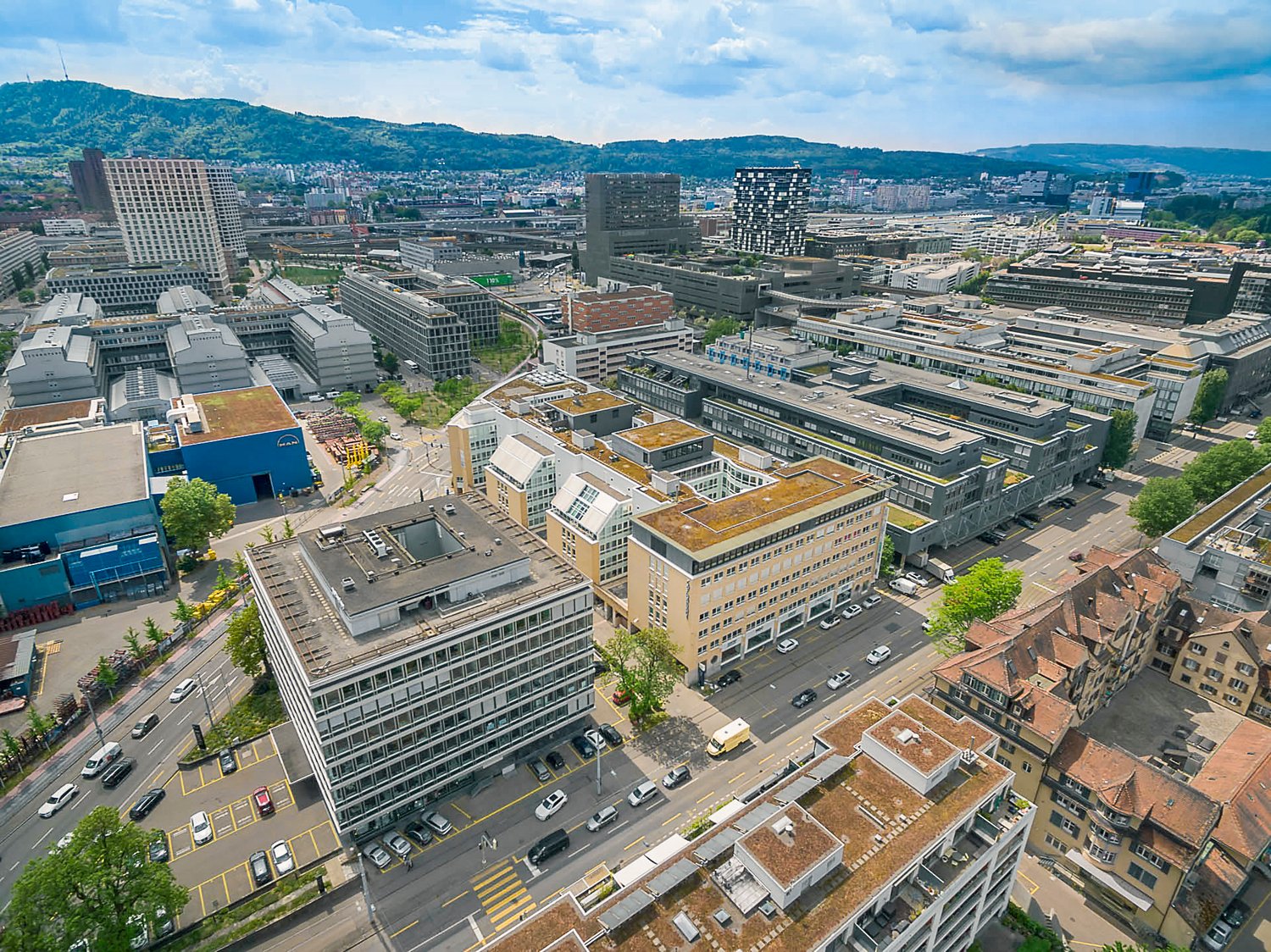 The image shows an aerial view of a dense urban cityscape with a mix of modern high-rise buildings and older structures. The city appears to be located in a hilly or mountainous area, with mountains visible in the background. The streets are lined with pa