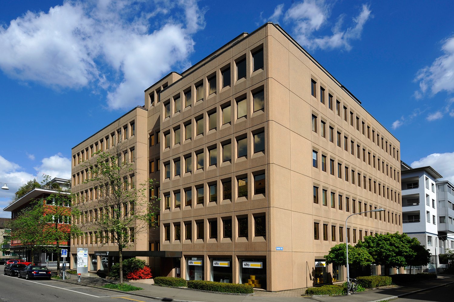 A multi-story office building with a beige facade, rectangular windows, and a modern architectural style. The building appears to be located in an urban setting with other buildings and greenery surrounding it.