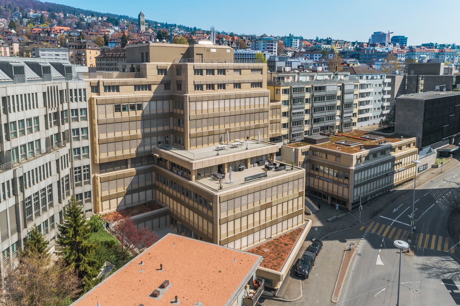 High rise office building with multiple floors, surrounded by urban landscape, several other buildings, green roofs, roads and vehicles