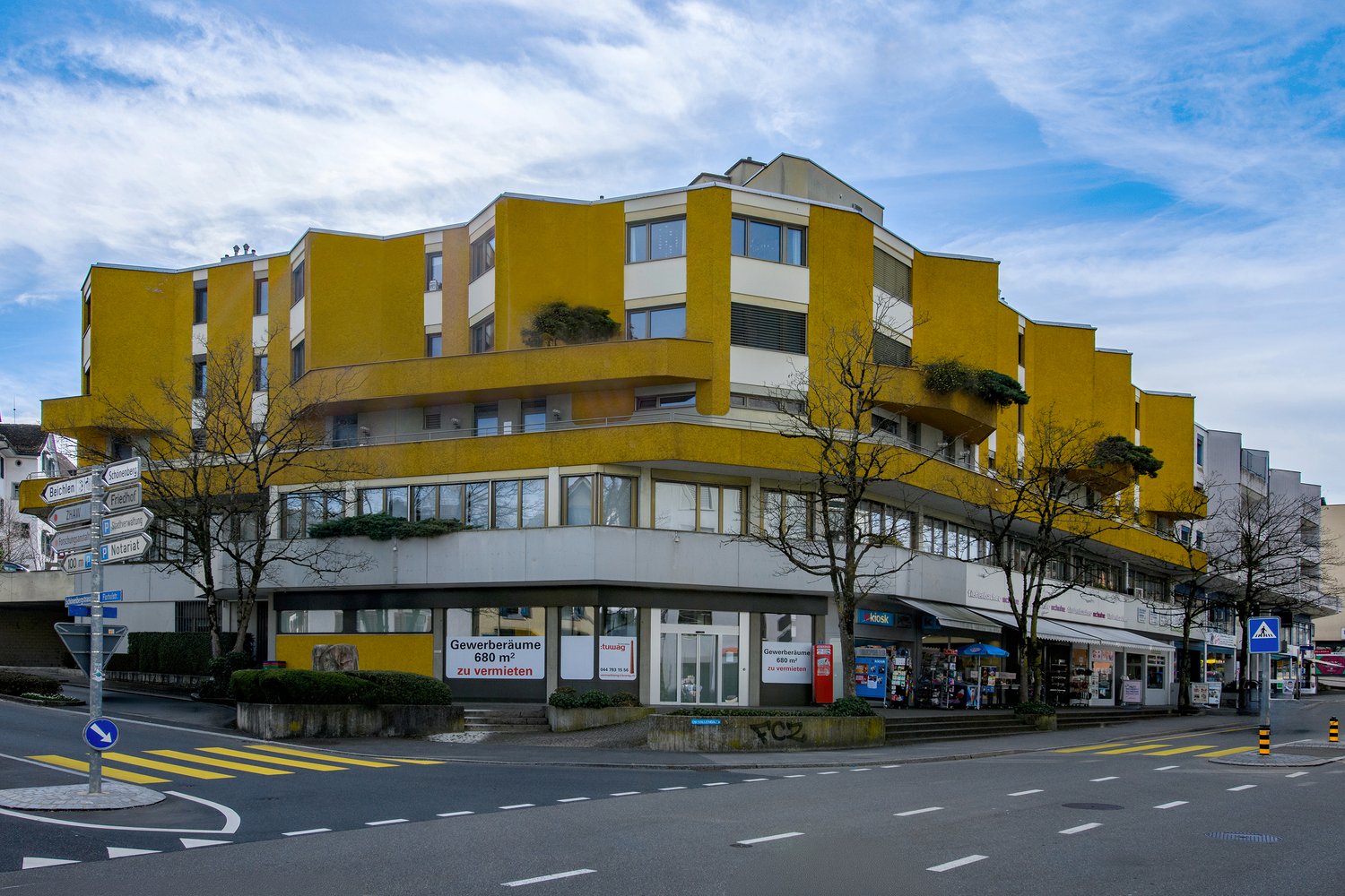 A yellow and white multi-story building with glass windows and balconies, located on a street with shops and trees.