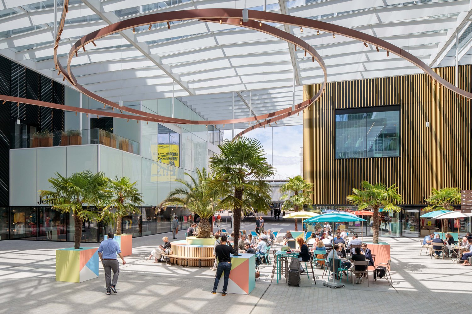 Large indoor commercial space with glass walls, curved copper-colored overhead structures, palm trees, outdoor seating area with tables and umbrellas, and people walking around and sitting at the tables.