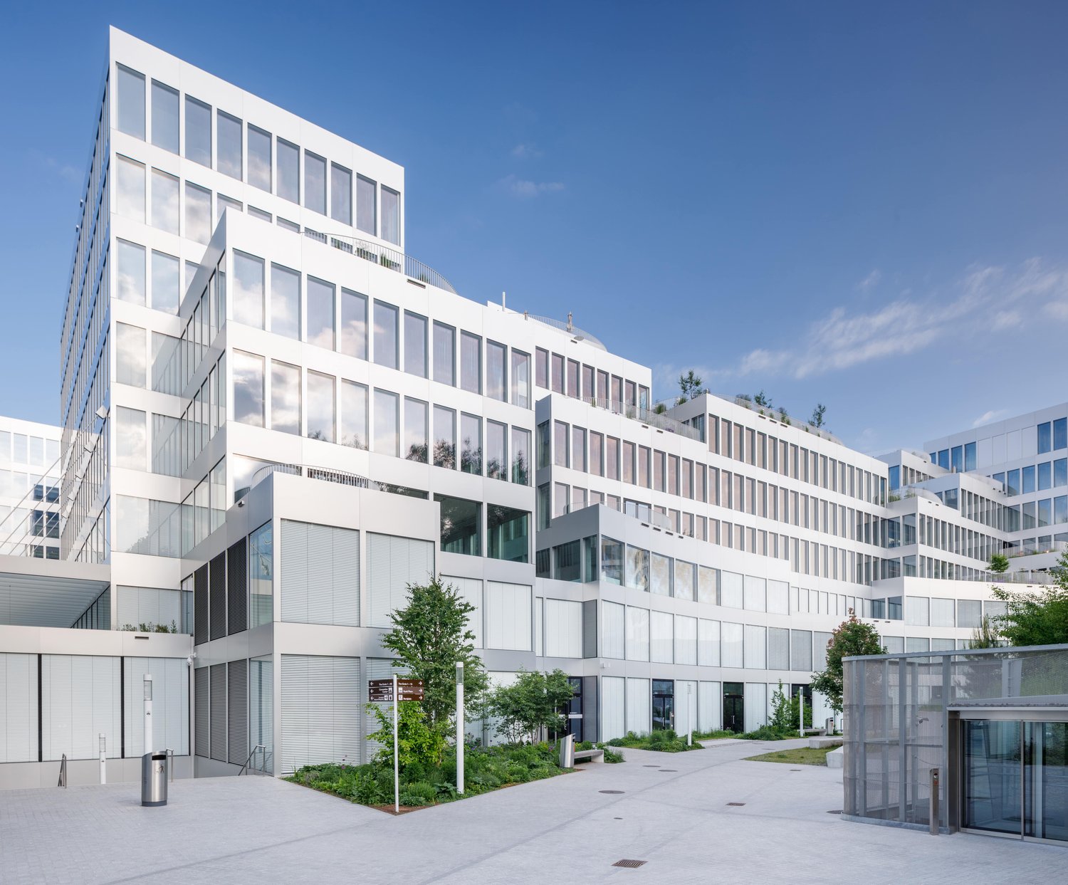 Multi-story office building with a modern, minimalist design featuring large windows, white exterior, and a landscaped courtyard with trees and greenery. The building appears to have a parking area or garage visible in the foreground.