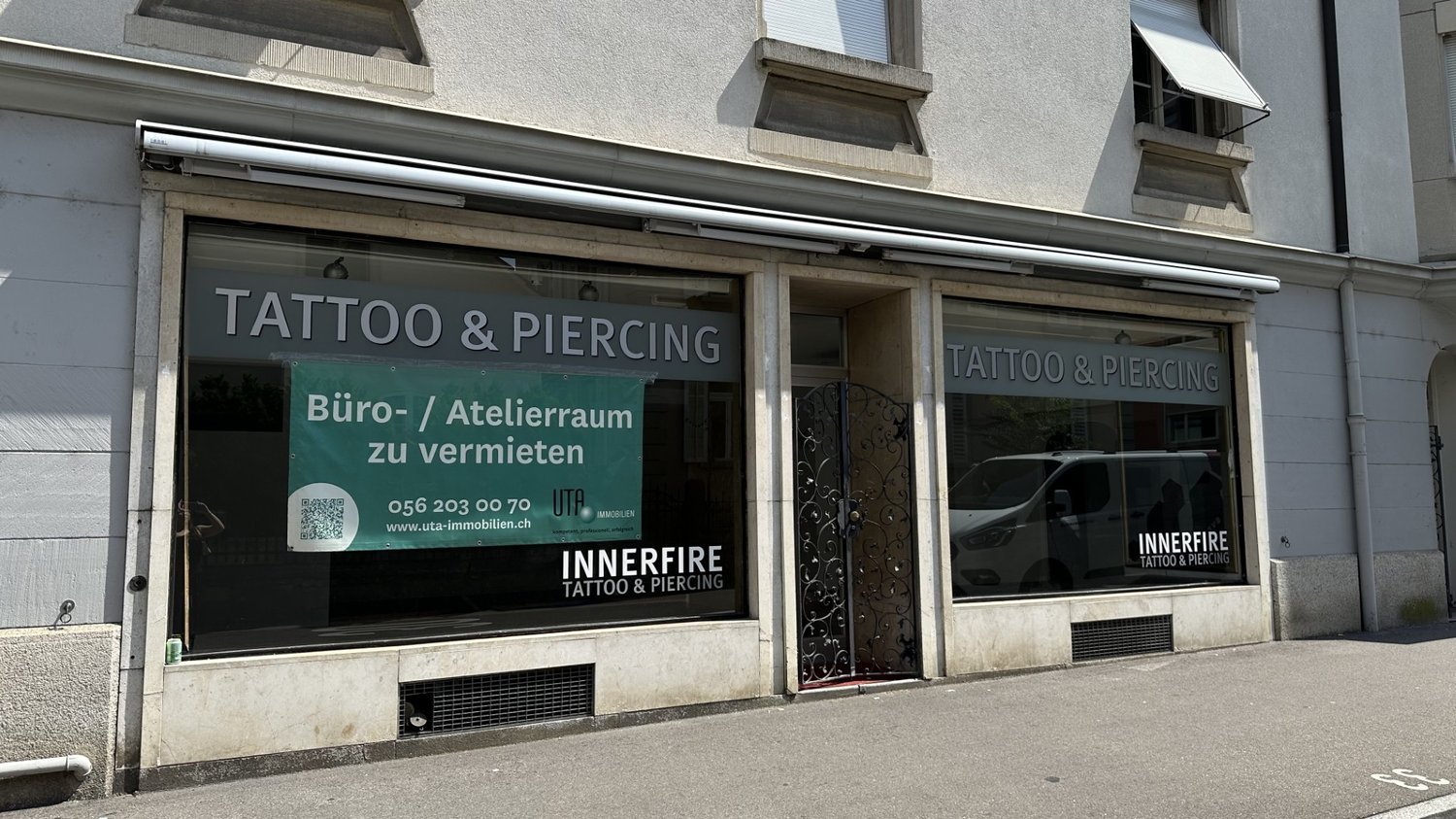 Storefront for tattoo and piercing studio with sign, windows and door visible.
