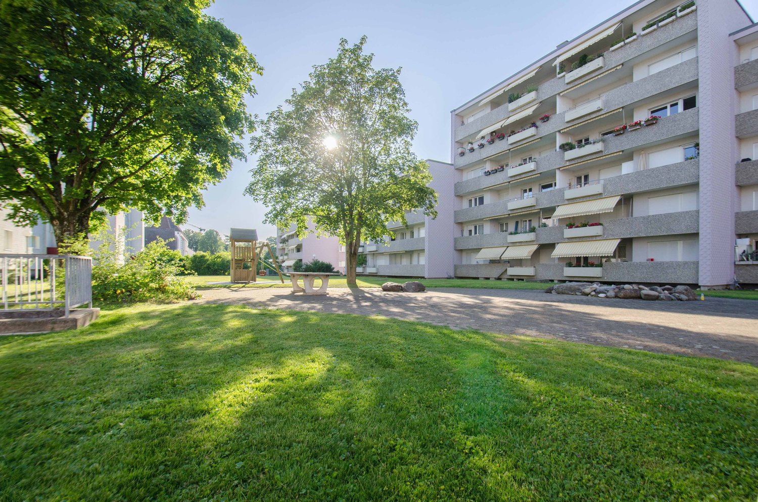 A grassy area with two trees and a playground, with a pink apartment building in the background.