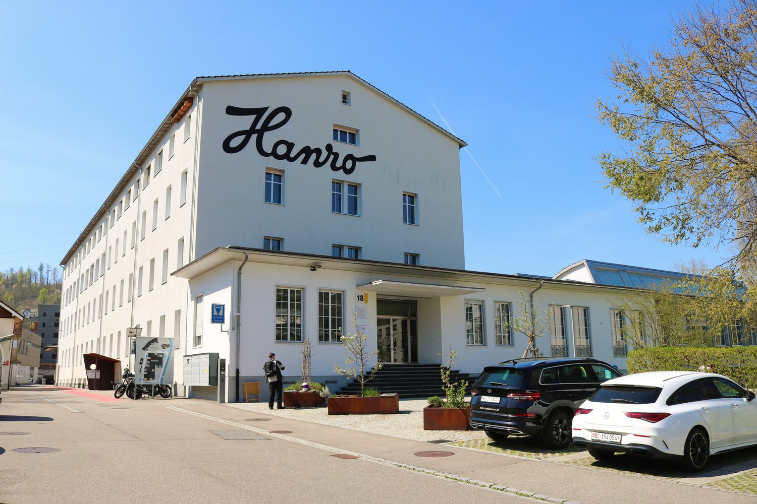 2-story white building, large glass windows, balcony on the second floor, cars parked in front, trees, bushes, walkway with a pedestrian