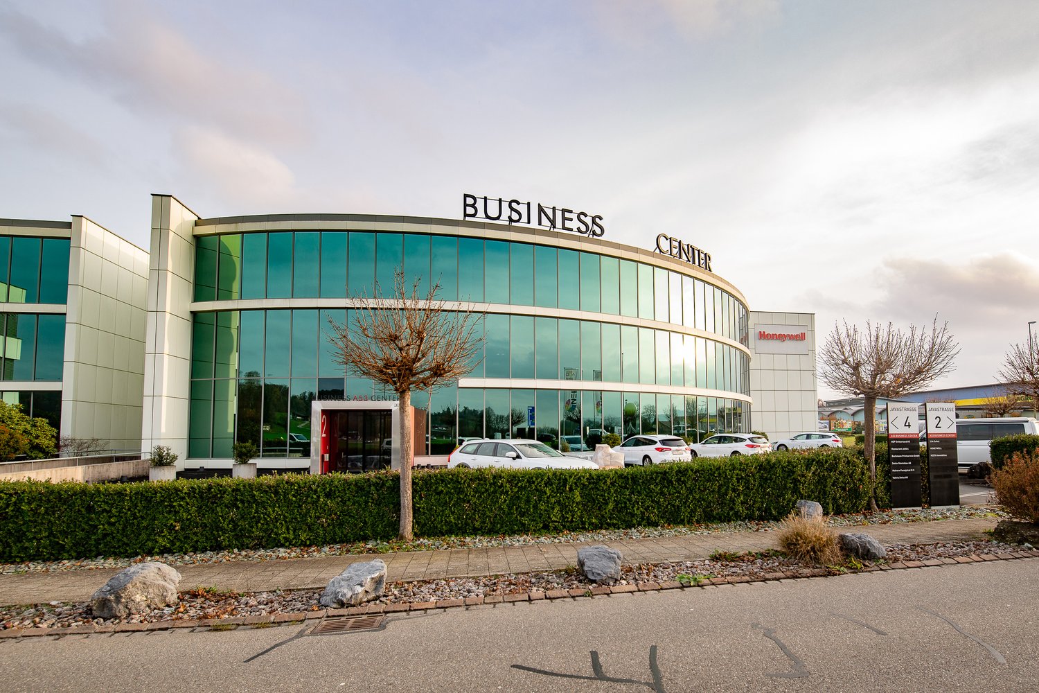 A large, multi-story glass and concrete building with the text 'BUSINESS CENTER' prominently displayed. The building has a curved facade and is surrounded by a parking lot with several cars parked. There is landscaping with trees and shrubs around the bui