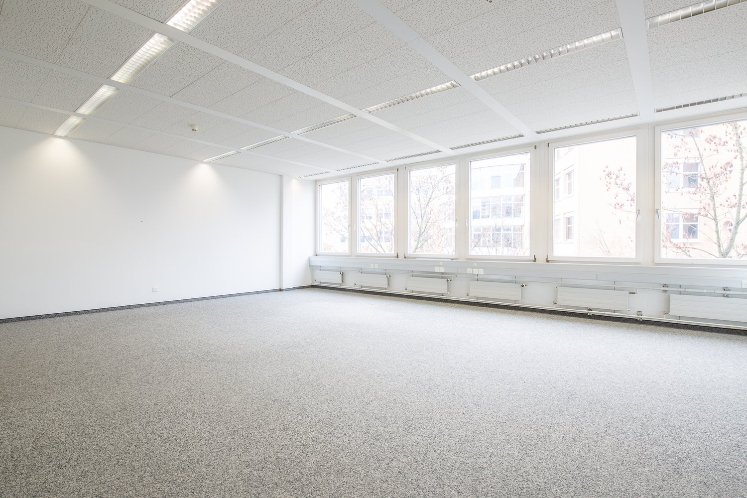 Large open office space with multiple windows providing a view, white walls, gray flooring, suspended ceiling with recessed lighting
