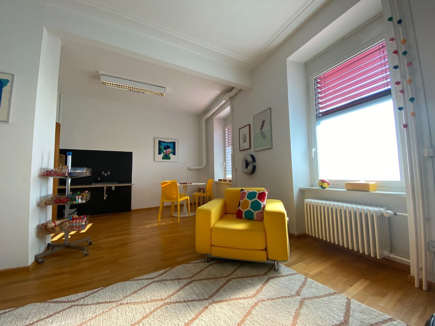 Bright and colorful living room with hardwood floors, white walls, large window, yellow armchair, shelving unit with toys and decor, and a patterned rug on the floor.