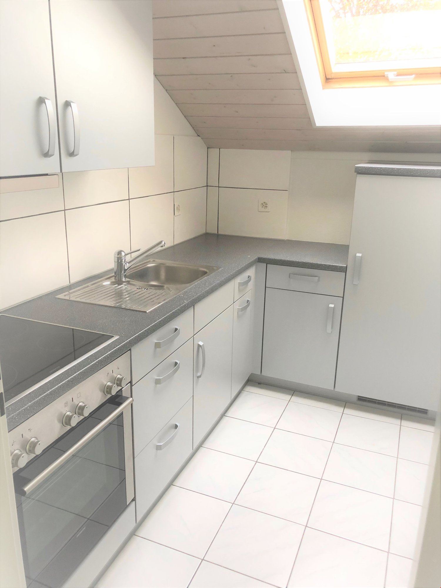 Compact kitchen with white cabinets, gray countertops, stainless steel sink, and built-in dishwasher. Tiled floor and slanted ceiling with a window.