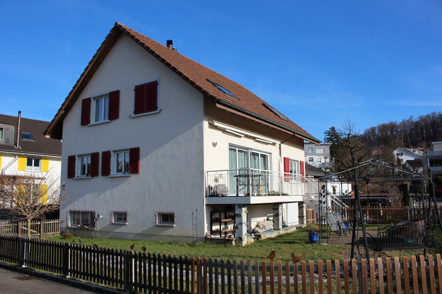 two-story house, white exterior, red shutters, brown roof, balcony with a railing, staircase, swing, garden with wooden fence, yellow house in the background