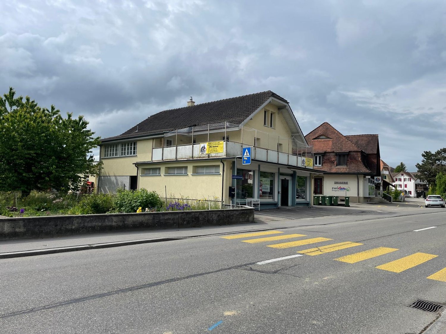 A two-story commercial building with a yellow exterior, a tiled roof, and several windows. The building has a grocery store called 'Swissco' on the ground floor. There is a crosswalk in front of the building and the surrounding area appears to be a reside
