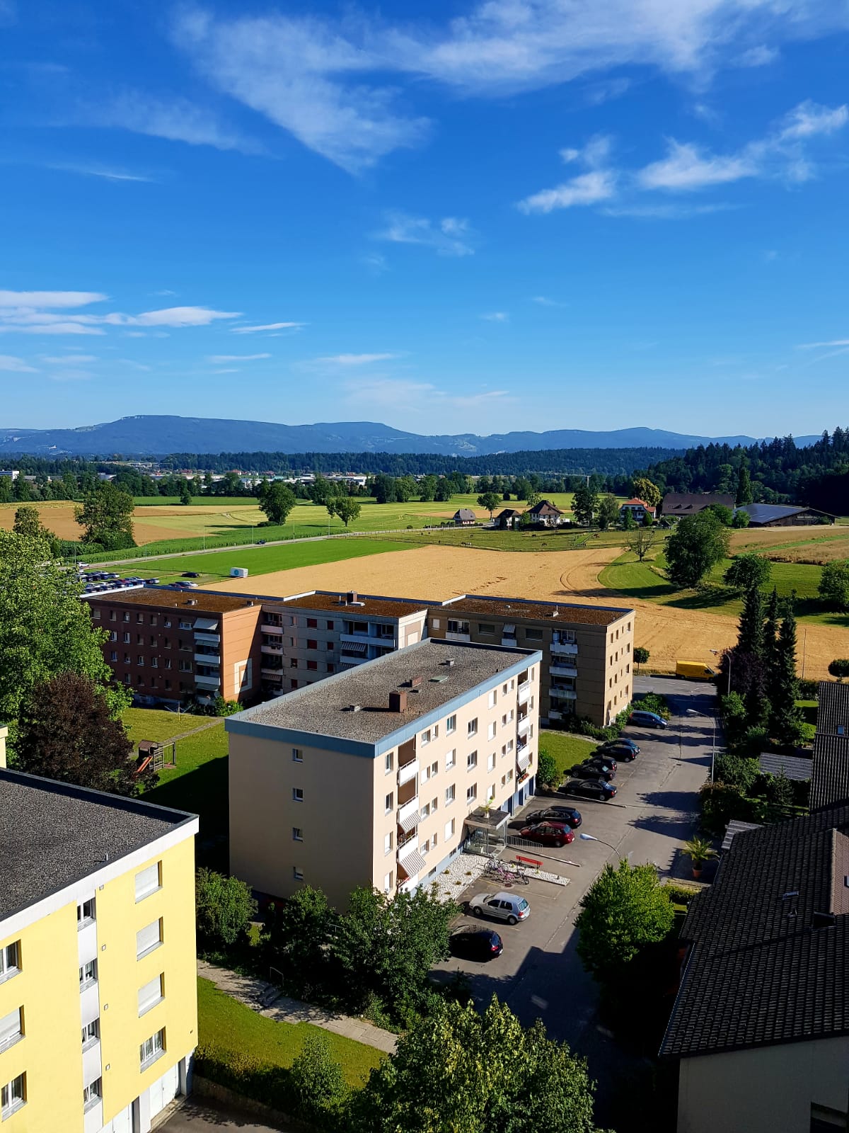 Multiple apartment buildings, large parking lot, trees, fields