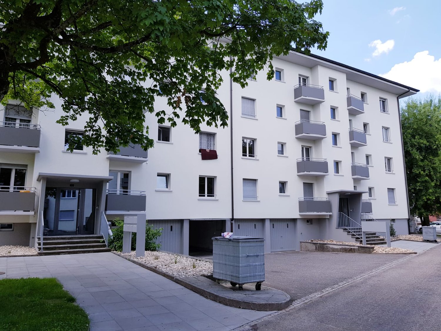 White multi-storey apartment building with multiple balconies, garage doors, entrance, steps, and paved parking area