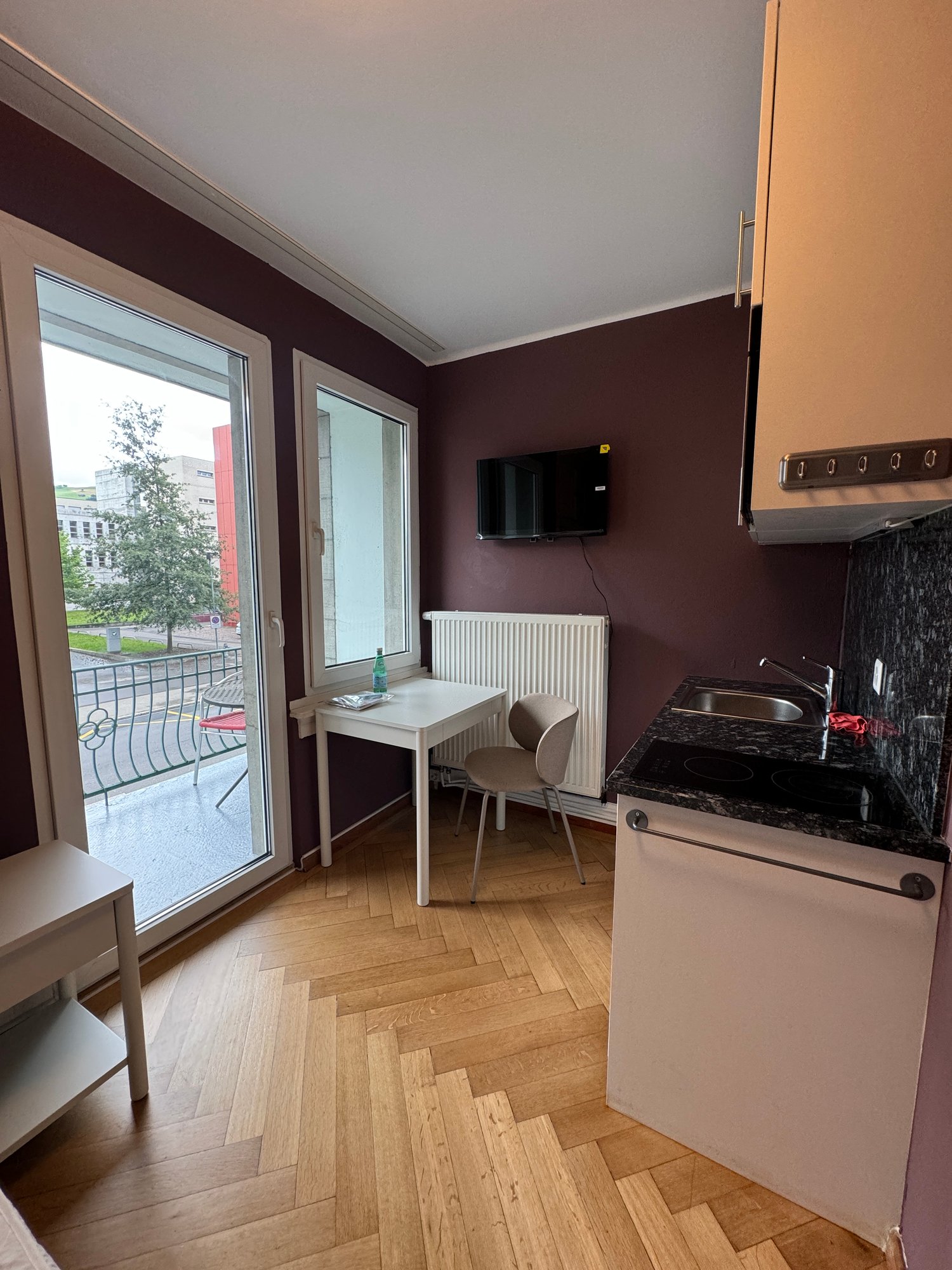 small room with a table, chair, and a television, a kitchen with a sink, stove, and a small refrigerator, a balcony with a view of the outside