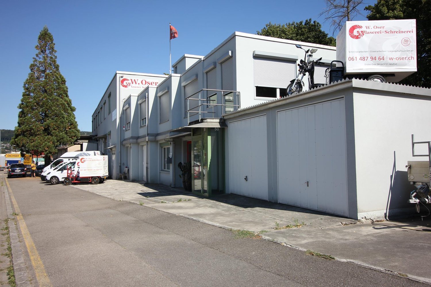 White industrial building, flag on the roof, roll-up doors, garage door, vehicles parked in front, trees and mountains in the background