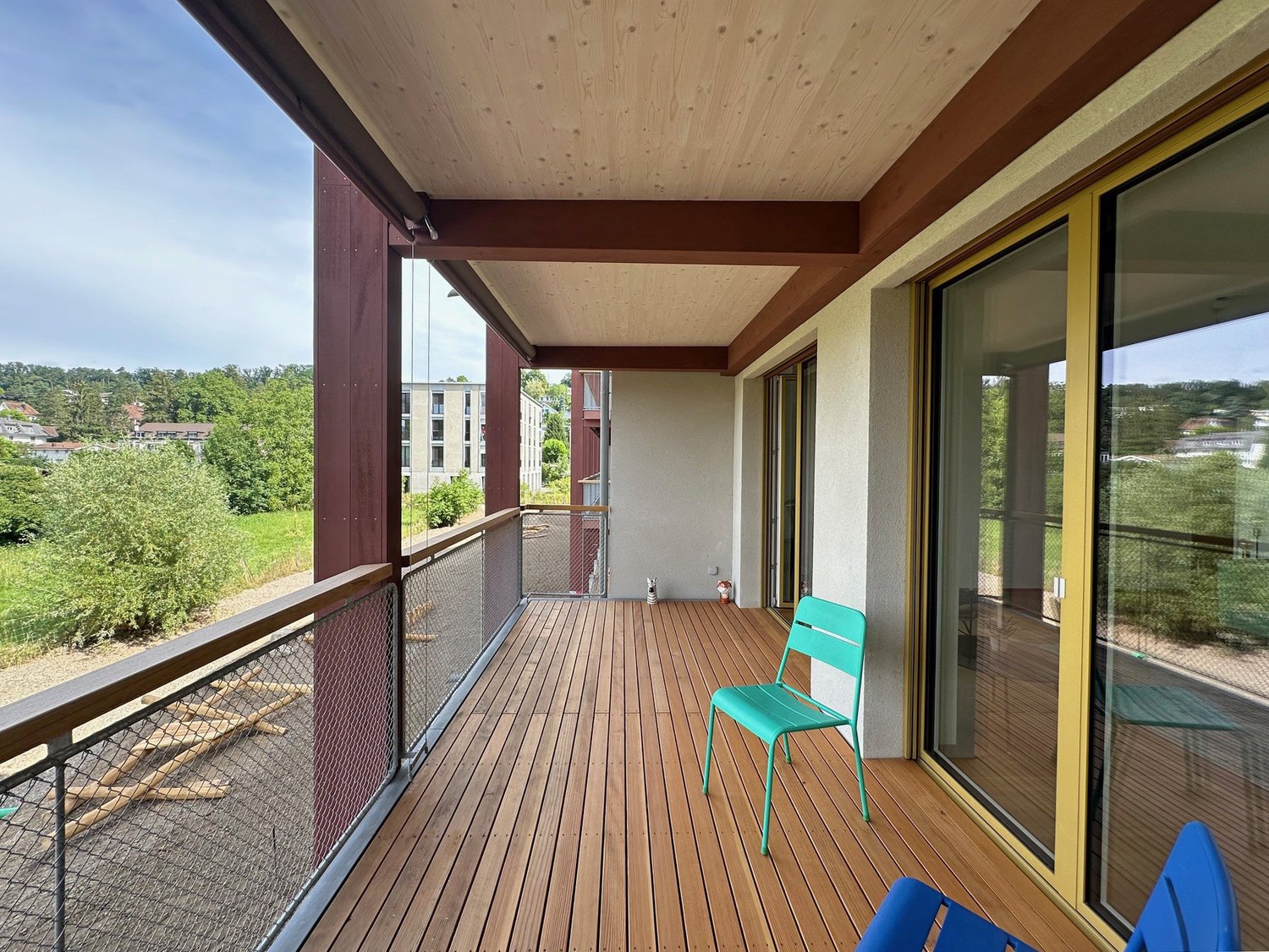 Wooden deck balcony with glass railing, overlooking a green, wooded area. The balcony has a turquoise chair and appears to be part of a larger residential building.