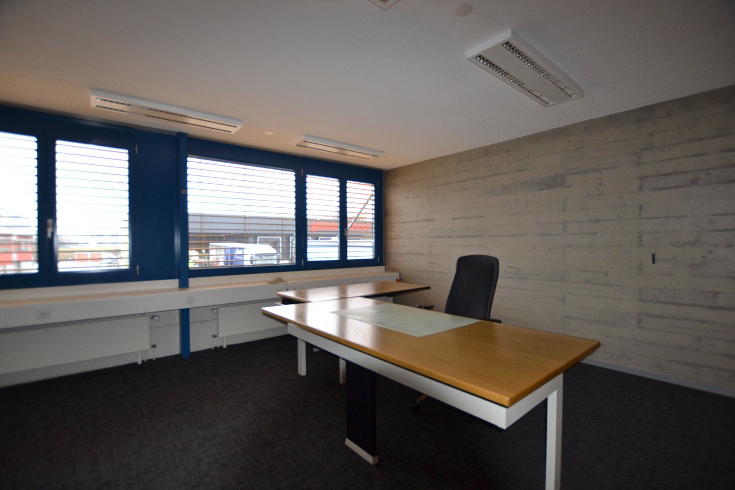 The image shows a modern office space with large windows, concrete walls, and wooden desks. The room has a minimalist and professional design, with a black office chair and multiple workstations set up for use.