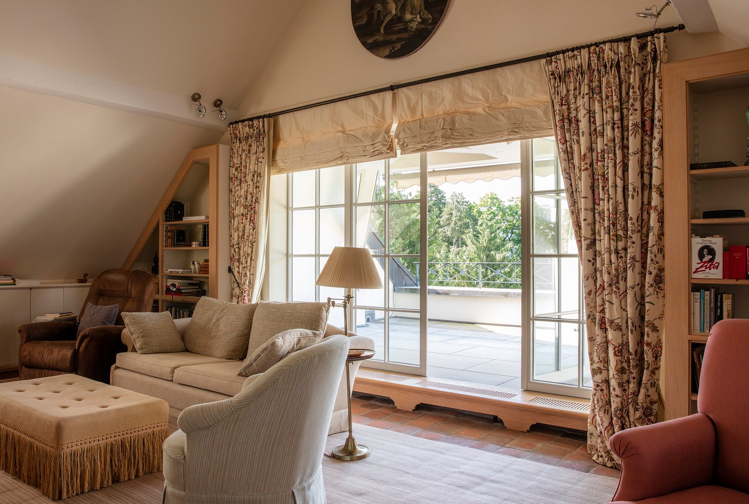 Cozy living room with large windows overlooking a balcony or terrace, beige sofa and armchair, wooden shelves, and a decorative painting on the wall