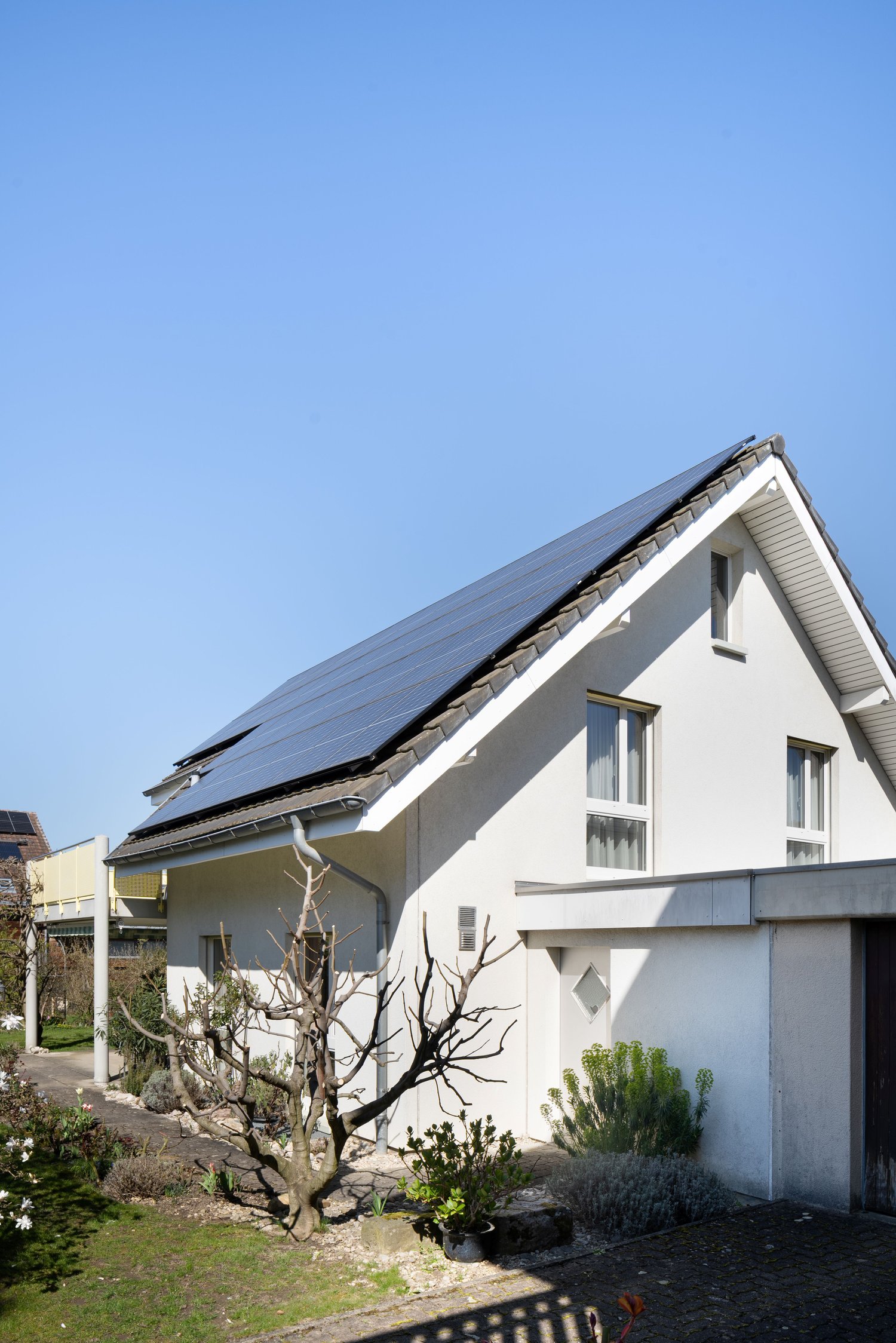 white house, solar panels on the roof, small garden, brick driveway