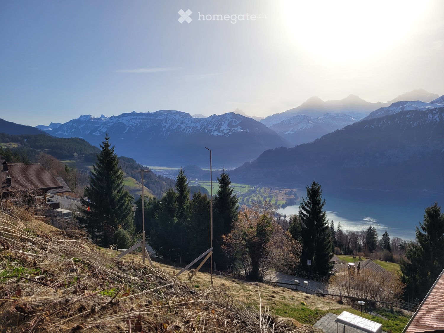 Aerial view of mountainous terrain with lush vegetation, a serene body of water, distant houses, and a clear sky.
