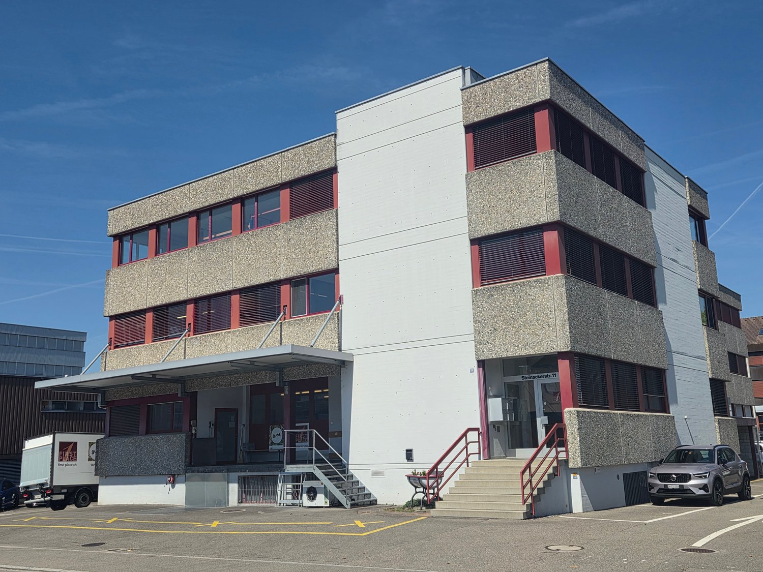 3-story commercial building, modern architectural design, multiple windows, stairs leading to the entrance, parking lot in front, adjacent building in the background