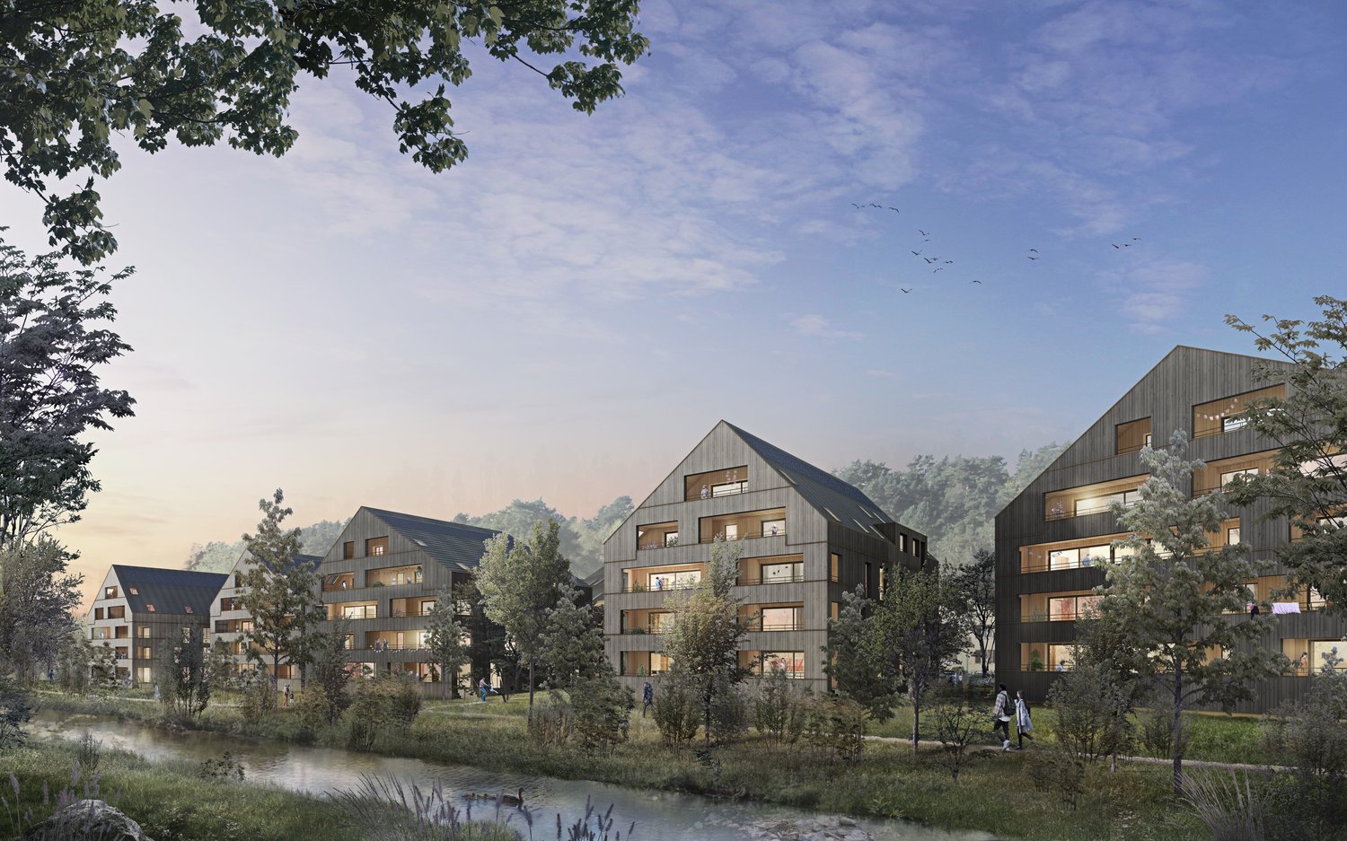 The image shows a residential complex with several multi-story buildings surrounded by trees and a small pond. The buildings have a modern, wooden-clad design with balconies visible on some of the units.