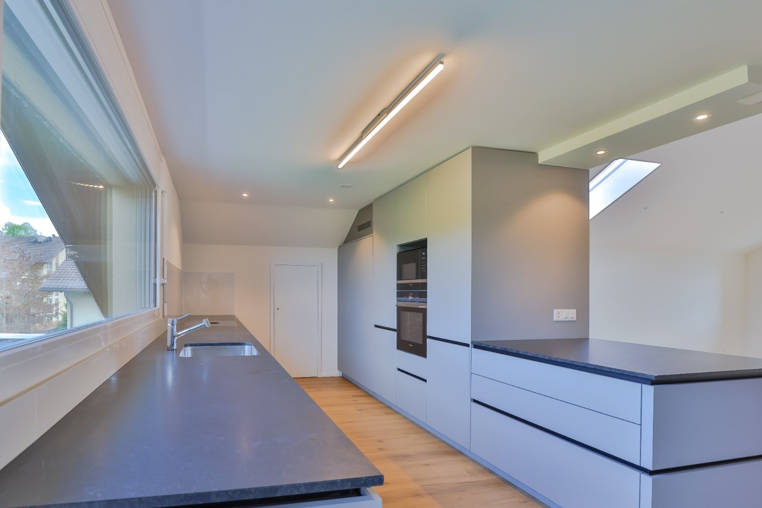 Modern kitchen with light grey cabinets, dark countertops, large window, embedded oven, double sinks, wooden floor.